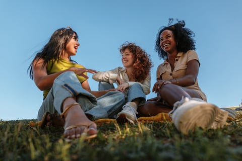 Three diverse female friends sitting on a blanket in a park laughing and enjoying each other's company on a sunny day