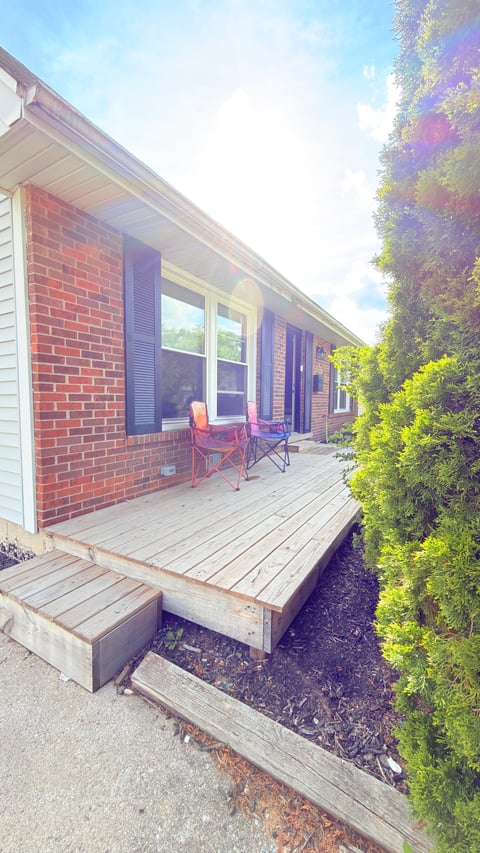 Brick house patio with wooden deck, two red chairs, large windows, blue shutters, and lush green vines along the right side